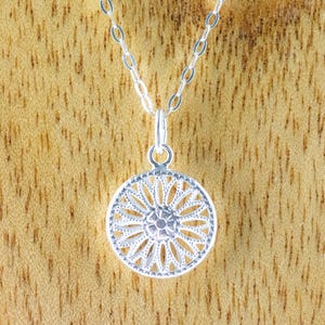 K&ouml;nnte beinhalten: Eine silberne Halskette mit einem zarten, runden Anh&auml;nger, der mit einem detaillierten Blumenmuster verziert ist. Der Anh&auml;nger erinnert an ein Mandala oder eine Sonnenstrahlen.
