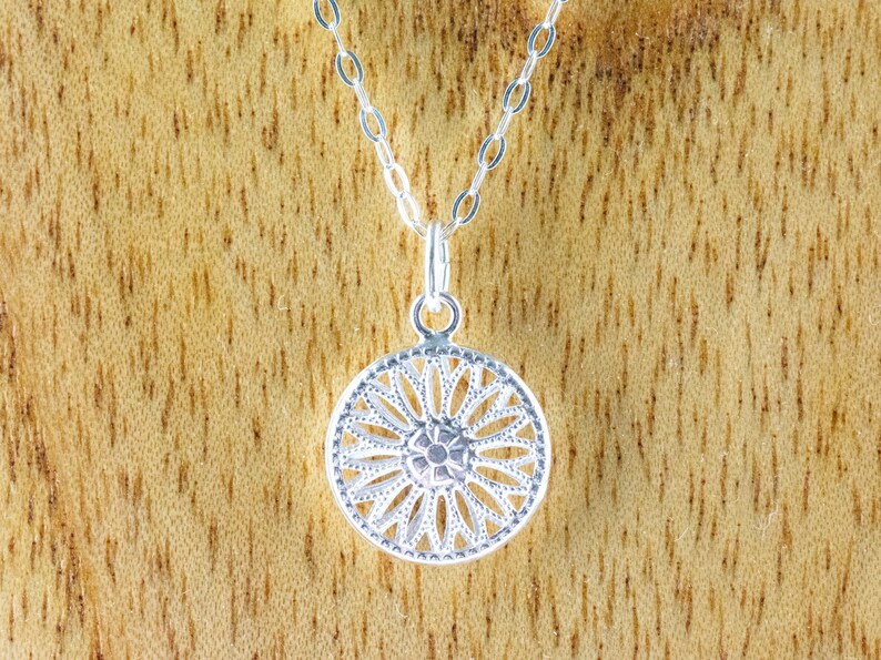 K&ouml;nnte beinhalten: Eine silberne Halskette mit einem zarten, runden Anh&auml;nger, der mit einem detaillierten Blumenmuster verziert ist. Der Anh&auml;nger erinnert an ein Mandala oder eine Sonnenstrahlen.