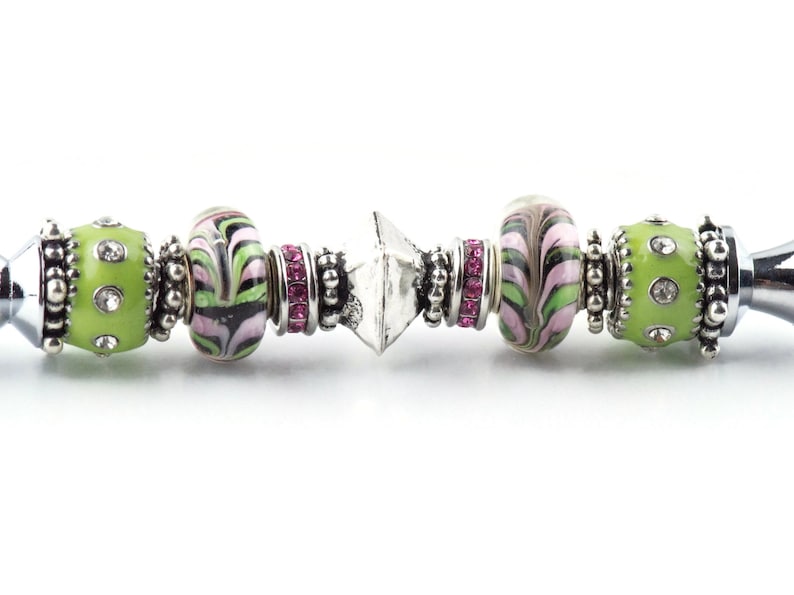 Peut inclure: Un bracelet &agrave; breloques argent&eacute; avec des perles vertes, roses et blanches. Les perles sont d&eacute;cor&eacute;es de volutes et de strass.
