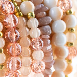 May include: A close-up of several strands of beaded bracelets. The bracelets are made with various shades of pink, peach, and white beads. Some beads are round, while others are oval or irregular in shape.