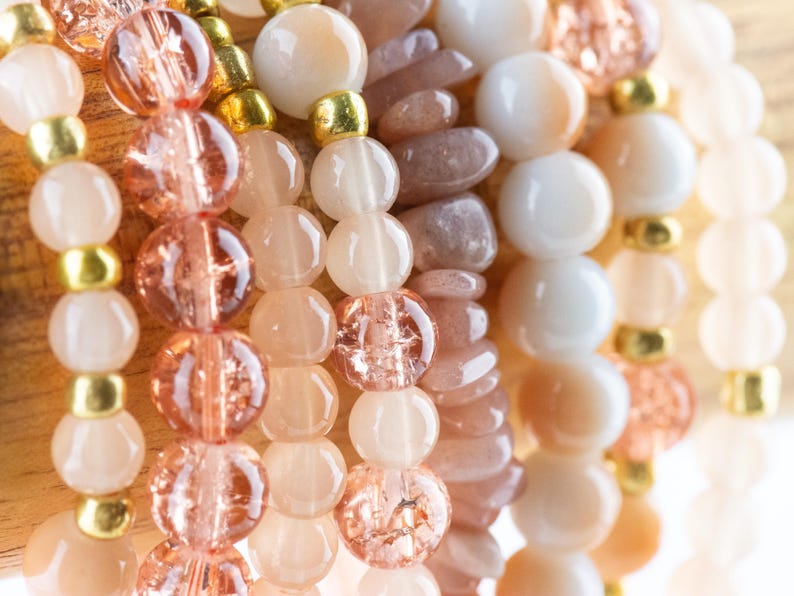 May include: A close-up of several strands of beaded bracelets. The bracelets are made with various shades of pink, peach, and white beads. Some beads are round, while others are oval or irregular in shape.