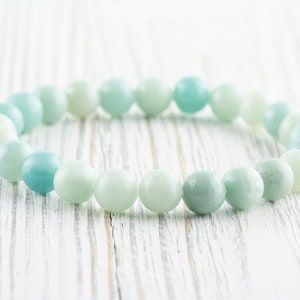 May include: A light blue and green beaded bracelet made of smooth, round stones. The bracelet is lying on a white wooden surface.
