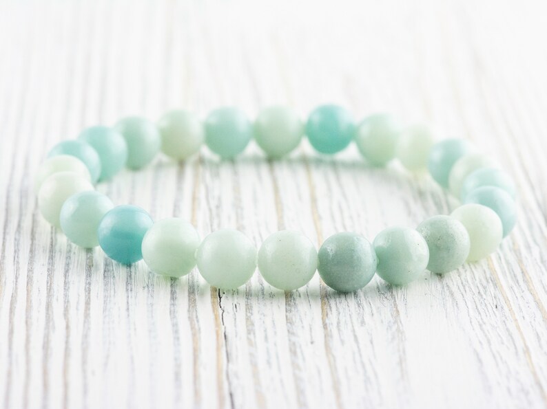 May include: A light blue and green beaded bracelet made of smooth, round stones. The bracelet is lying on a white wooden surface.