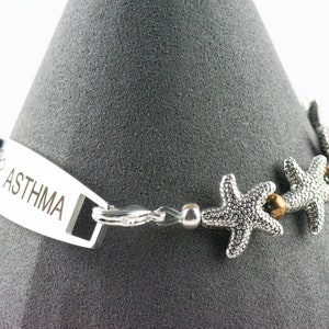 K&ouml;nnte beinhalten: Ein silbernes medizinisches Armband mit der Gravur "ASTHMA". Das Armband hat einen silbernen Verschluss und ist mit silbernen Seestern-Anh&auml;ngern verziert.