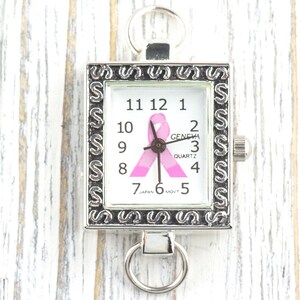 Op de afbeelding: Zilverkleurig vierkant horloge met een witte wijzerplaat met zwarte cijfers en een roze lint. De wijzerplaat toont "GENEVA QUARTZ" en "JAPAN MOVT". Het heeft een decoratieve rand en een ring aan de boven- en onderkant.