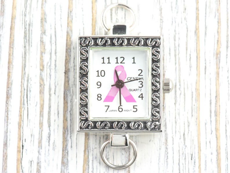 Op de afbeelding: Zilverkleurig vierkant horloge met een witte wijzerplaat met zwarte cijfers en een roze lint. De wijzerplaat toont "GENEVA QUARTZ" en "JAPAN MOVT". Het heeft een decoratieve rand en een ring aan de boven- en onderkant.