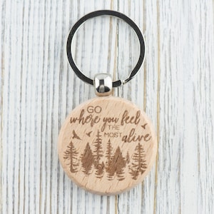 May include: Wooden keychain with a round, light brown tag featuring a forest scene and the text "Go where you feel the most alive".