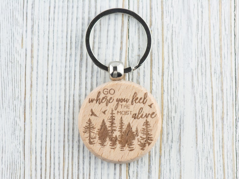 May include: Wooden keychain with a round, light brown tag featuring a forest scene and the text "Go where you feel the most alive".