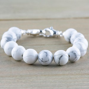 Peut inclure: Un bracelet en perles d'howlite blanches avec un fermoir argent&eacute;. Les perles sont rondes et ont un motif marbr&eacute;.