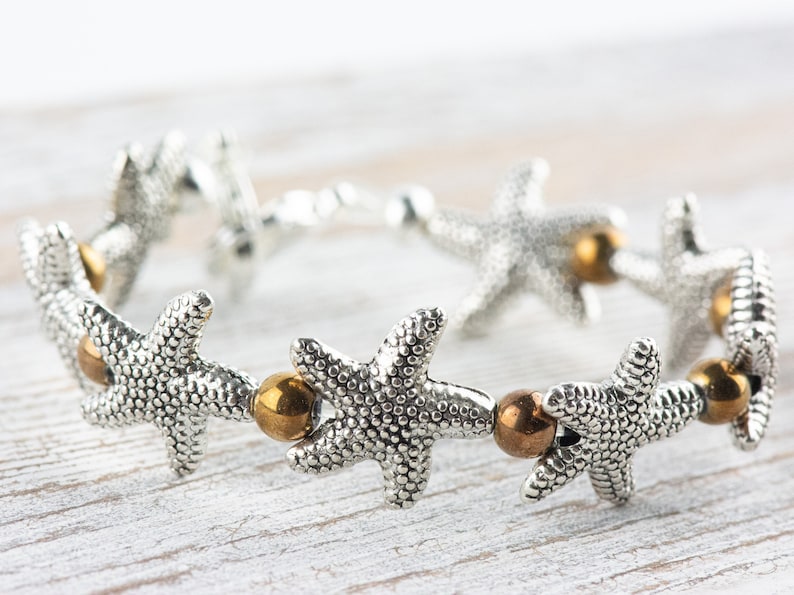 K&ouml;nnte beinhalten: Silberarmband mit einer Reihe von Seestern-Anh&auml;ngern und goldfarbenen Perlen.