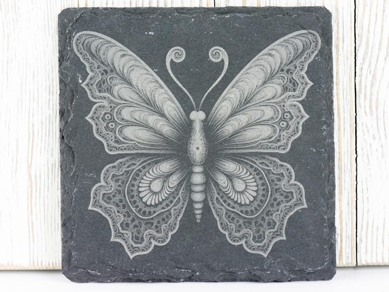 Puede incluir: Un posavasos de pizarra negra con un dise&ntilde;o de mariposa blanca intrincada grabado en la superficie.