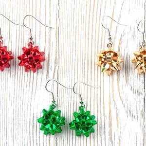 May include: Three pairs of festive bow earrings in red, gold, and green. Each earring features a bow design with a silver hook. The earrings are displayed on a white wooden surface, perfect for holiday or special occasion wear.