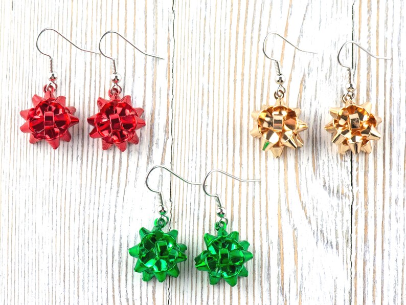 May include: Three pairs of festive bow earrings in red, gold, and green. Each earring features a bow design with a silver hook. The earrings are displayed on a white wooden surface, perfect for holiday or special occasion wear.