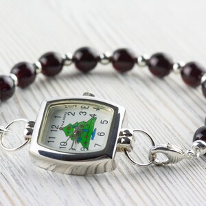 May include: A silver-toned watch with a square face and a Christmas tree design on the dial. The watch is attached to a bracelet made of dark red beads and silver-toned spacers. The watch face reads "NARMI".