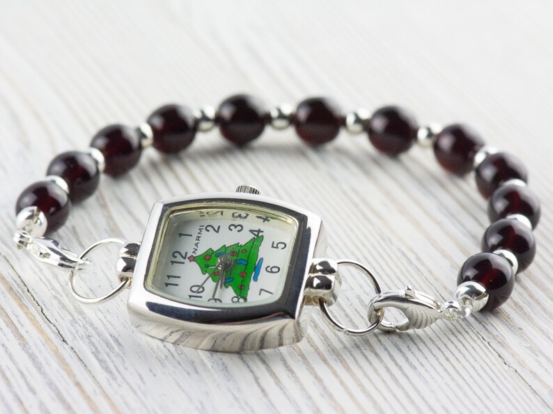 May include: A silver-toned watch with a square face and a Christmas tree design on the dial. The watch is attached to a bracelet made of dark red beads and silver-toned spacers. The watch face reads "NARMI".