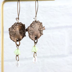May include: A pair of vintage-style earrings. Each earring features a bronze-colored, circular pendant with a bird and floral design. Below the pendant hang a light green flower and a clear teardrop-shaped bead. The earrings are suspended from a gold-colored hook.