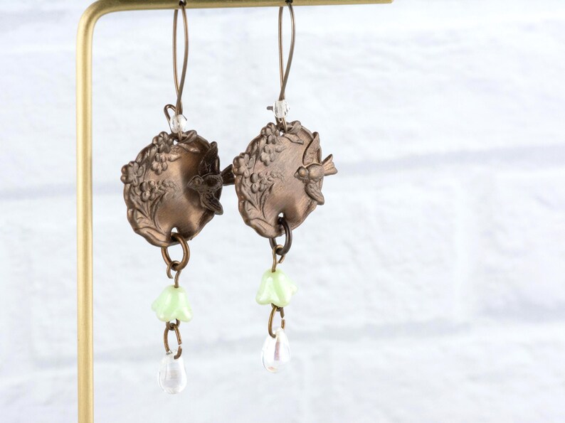 May include: A pair of vintage-style earrings. Each earring features a bronze-colored, circular pendant with a bird and floral design. Below the pendant hang a light green flower and a clear teardrop-shaped bead. The earrings are suspended from a gold-colored hook.