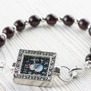 K&ouml;nnte beinhalten: Eine silberne Uhr mit schwarzem Zifferblatt und einem Schneemann-Motiv. Die Uhr ist an einem Armband aus dunkelroten Perlen befestigt.