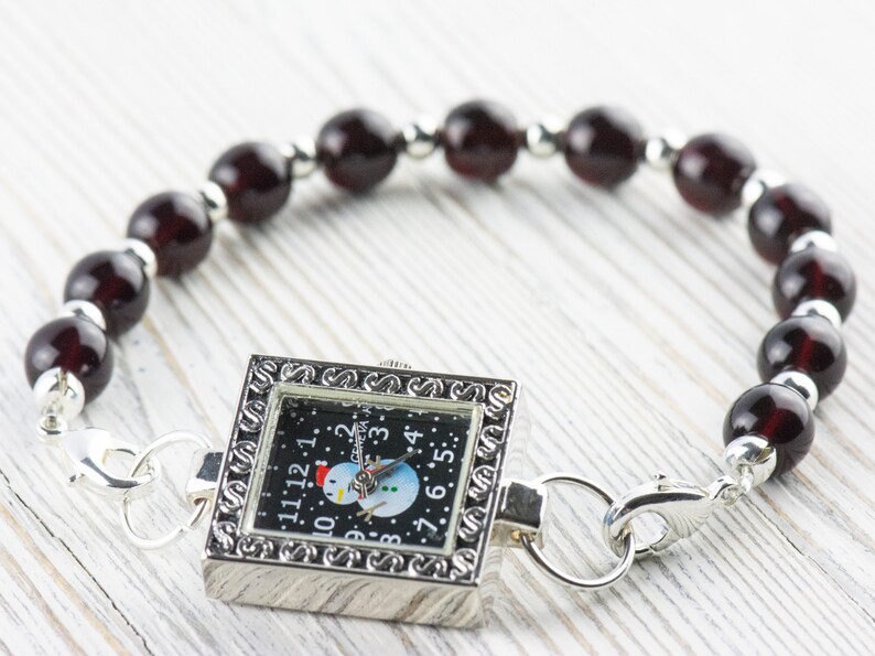 K&ouml;nnte beinhalten: Eine silberne Uhr mit schwarzem Zifferblatt und einem Schneemann-Motiv. Die Uhr ist an einem Armband aus dunkelroten Perlen befestigt.