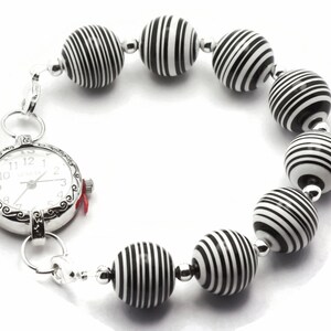 K&ouml;nnte beinhalten: Eine silberne Armbanduhr mit wei&szlig;em Zifferblatt und schwarzen Zahlen ist an einem Armband aus schwarz-wei&szlig; gestreiften Perlen befestigt.