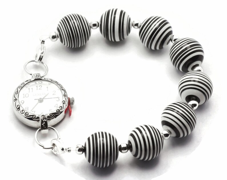 K&ouml;nnte beinhalten: Eine silberne Armbanduhr mit wei&szlig;em Zifferblatt und schwarzen Zahlen ist an einem Armband aus schwarz-wei&szlig; gestreiften Perlen befestigt.