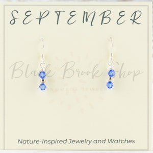 May include: A pair of silver earrings with a blue crystal charm and a gold bead. The earrings are displayed on a card that says "SEPTEMBER" and "Black Brook Shop".