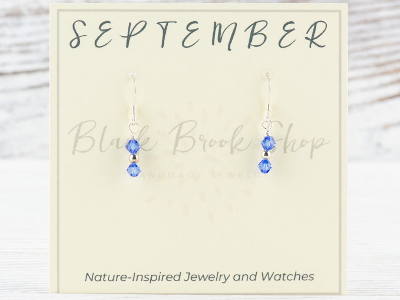May include: A pair of silver earrings with a blue crystal charm and a gold bead. The earrings are displayed on a card that says "SEPTEMBER" and "Black Brook Shop".