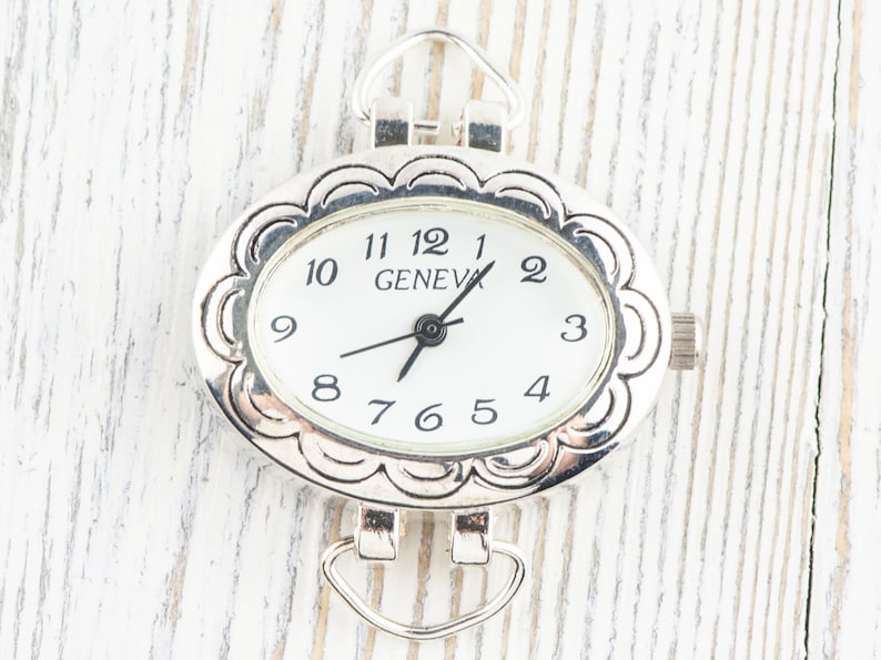 May include: Silver-coloured oval watch face with a white dial and black numerals. The watch face has a decorative scalloped edge and the word "GENEVA" is printed on the dial. It has loops for attaching to a chain or strap.