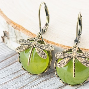 May include: A pair of green glass and bronze dragonfly earrings. The earrings feature a round green glass cabochon with a bronze dragonfly design on top. The earrings are attached to bronze leverback ear wires.