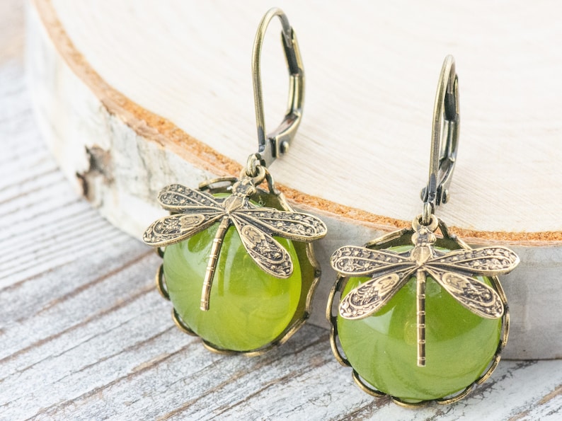 May include: A pair of green glass and bronze dragonfly earrings. The earrings feature a round green glass cabochon with a bronze dragonfly design on top. The earrings are attached to bronze leverback ear wires.