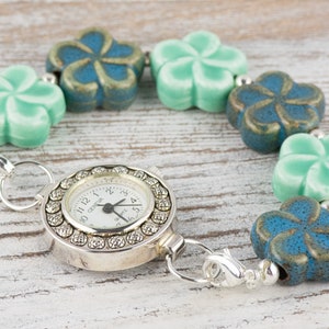 May include: A silver pocket watch with a white face and black numbers is attached to a bracelet made of alternating blue and green ceramic flower beads.