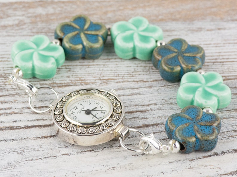 May include: A silver pocket watch with a white face and black numbers is attached to a bracelet made of alternating blue and green ceramic flower beads.