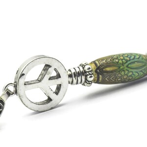 Puede incluir: Un collar con un colgante de plata en forma de signo de paz y una cuenta con un patr&oacute;n verde y marr&oacute;n. La cuenta tiene un dise&ntilde;o floral.
