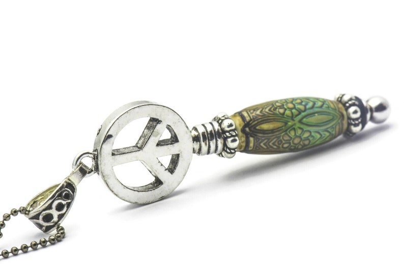 Puede incluir: Un collar con un colgante de plata en forma de signo de paz y una cuenta con un patr&oacute;n verde y marr&oacute;n. La cuenta tiene un dise&ntilde;o floral.