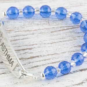 Pu&ograve; includere: Un braccialetto di allerta medica in argento con perline di vetro blu. Il braccialetto ha un'etichetta con la scritta "ASTHMA" con un simbolo di stella della vita.