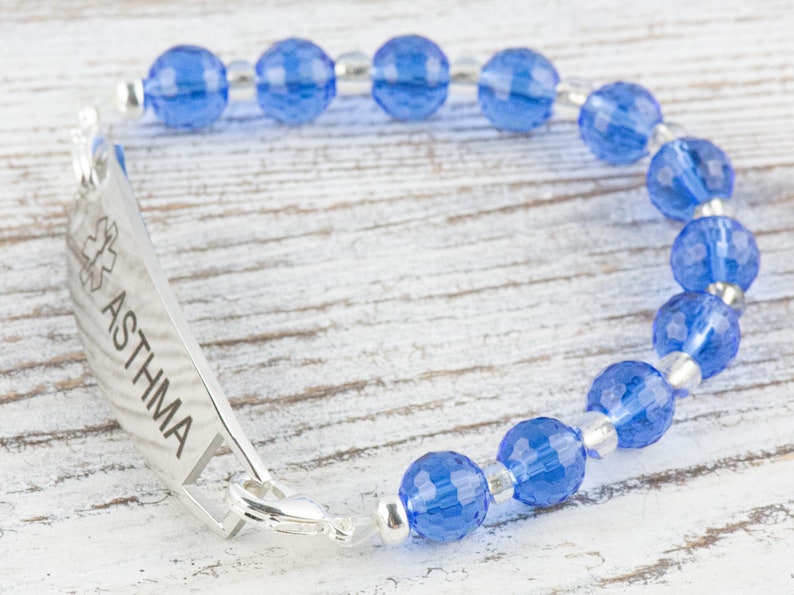 Pu&ograve; includere: Un braccialetto di allerta medica in argento con perline di vetro blu. Il braccialetto ha un'etichetta con la scritta "ASTHMA" con un simbolo di stella della vita.