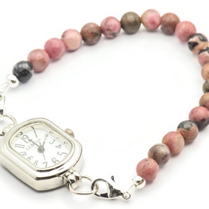 K&ouml;nnte beinhalten: Eine silberne Uhr mit quadratischem Zifferblatt und schwarzen r&ouml;mischen Ziffern an einem rosafarbenen und braunen Perlenarmband.