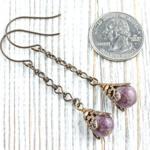May include: Handmade earrings with a purple gemstone and bronze chain. The earrings have a decorative cap and are shown next to a coin for size comparison. The earrings are approximately 6 cm in length.