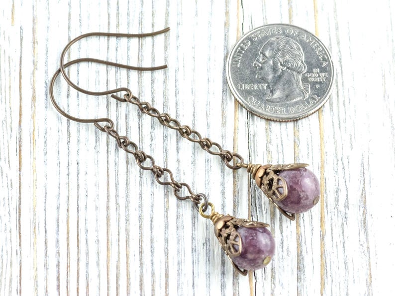 May include: Handmade earrings with a purple gemstone and bronze chain. The earrings have a decorative cap and are shown next to a coin for size comparison. The earrings are approximately 6 cm in length.