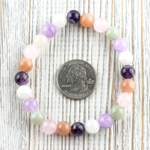 May include: A beaded bracelet featuring a variety of colorful gemstones, including purple, pink, peach, and white. The bracelet is circular, with a U.S. quarter coin in the center for size comparison. The beads are smooth and round, strung together on an elastic band.