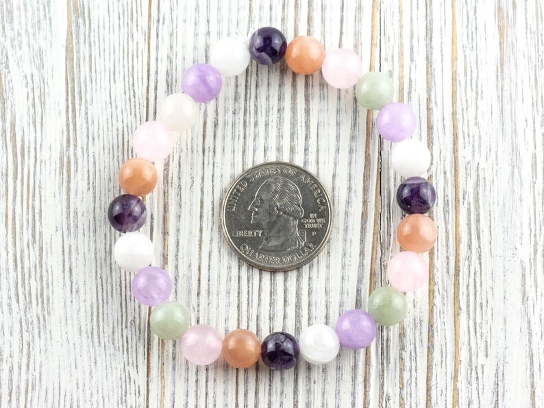 May include: A beaded bracelet featuring a variety of colorful gemstones, including purple, pink, peach, and white. The bracelet is circular, with a U.S. quarter coin in the center for size comparison. The beads are smooth and round, strung together on an elastic band.