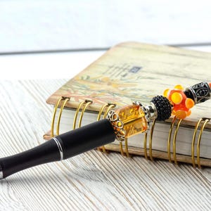 May include: A black pen with a decorative beaded top featuring orange, black, and silver accents. The pen rests on a spiral-bound notebook with a floral design, set against a white wooden surface.