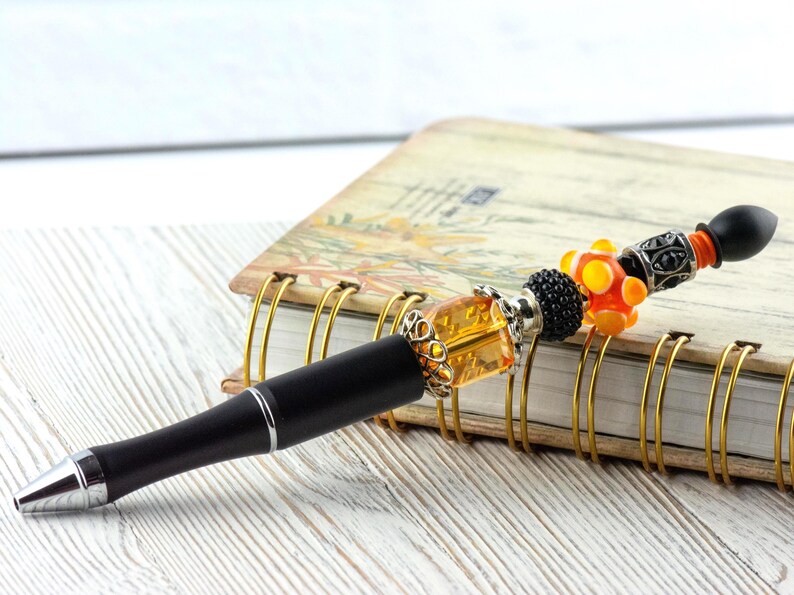 May include: A black pen with a decorative beaded top featuring orange, black, and silver accents. The pen rests on a spiral-bound notebook with a floral design, set against a white wooden surface.