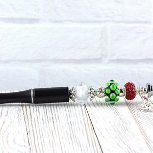 May include: A black pen with silver accents and decorative beads. The beads include a clear, a green with white dots, and a red bead. The pen has a black, textured barrel and a silver tip. A small charm hangs from the pen.