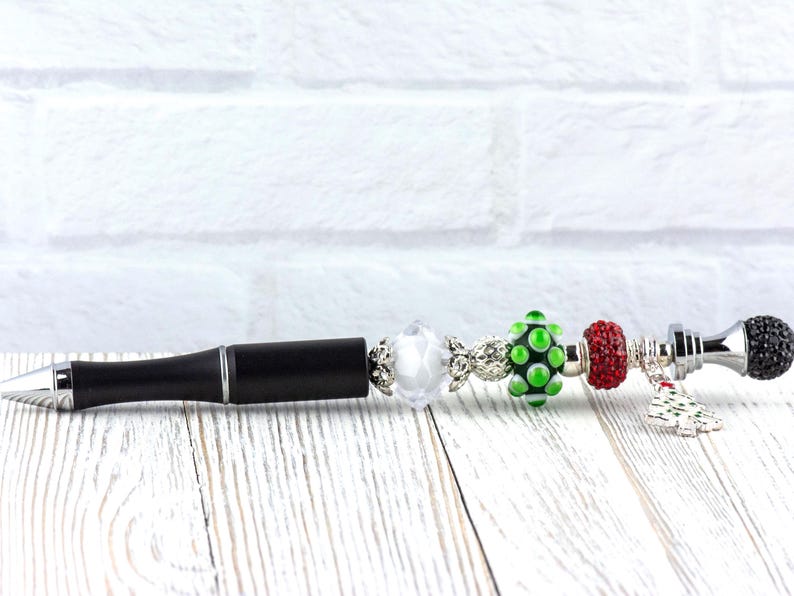 May include: A black pen with silver accents and decorative beads. The beads include a clear, a green with white dots, and a red bead. The pen has a black, textured barrel and a silver tip. A small charm hangs from the pen.