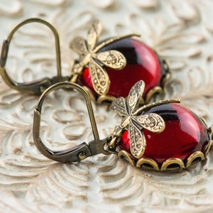 Pode incluir: Um par de brincos pendentes com caboch&otilde;es de vidro vermelho e detalhes de lib&eacute;lula dourados. Os brincos t&ecirc;m fecho de gancho.