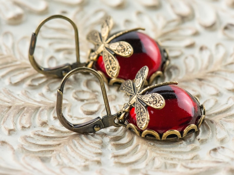 Pode incluir: Um par de brincos pendentes com caboch&otilde;es de vidro vermelho e detalhes de lib&eacute;lula dourados. Os brincos t&ecirc;m fecho de gancho.