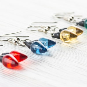 Op de afbeelding: Een set van vijf paar hangende oorbellen met lichtbollen van glas in de kleuren rood, blauw, geel, groen en helder. De oorbellen zijn gemaakt met zilverkleurige metalen haakjes.