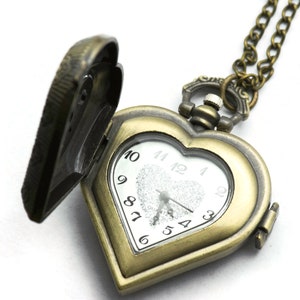 Puede incluir: Un reloj de bolsillo en forma de coraz&oacute;n de bronce antiguo con una tapa abierta, que revela una esfera blanca con n&uacute;meros negros. El reloj cuelga de una cadena a juego. La esfera del reloj tiene la palabra "QUARTZ" impresa.