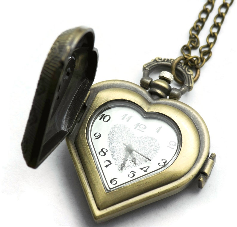 Puede incluir: Un reloj de bolsillo en forma de coraz&oacute;n de bronce antiguo con una tapa abierta, que revela una esfera blanca con n&uacute;meros negros. El reloj cuelga de una cadena a juego. La esfera del reloj tiene la palabra "QUARTZ" impresa.